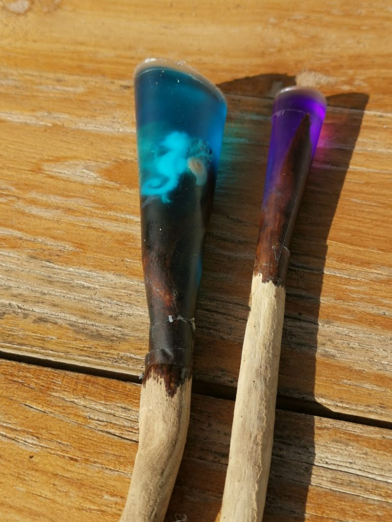 Twee drijfhout stokjes met UV-resin uiteinden in turquoise blauw en paars op een drijfhout achtergrond