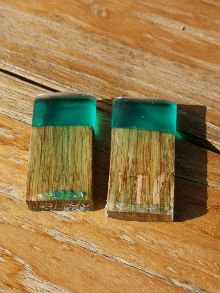 Twee kleine drijfhout blokjes gevat in smaragdgroen transparant epoxy op een houten ondergrond