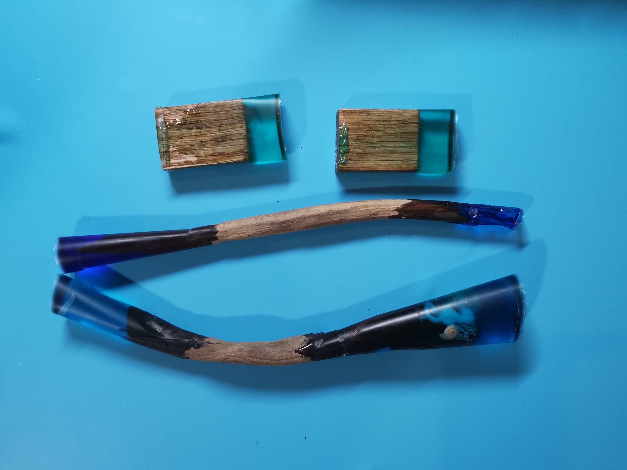 Alle stukken plat op een blauwe siliconen mat: eikblokjes met uitgeharde teal resin, drijfhout hairpins met uitgeharde dop aan de bovenkant en transparante resintape mallen gevuld met verse resin aan de punt
