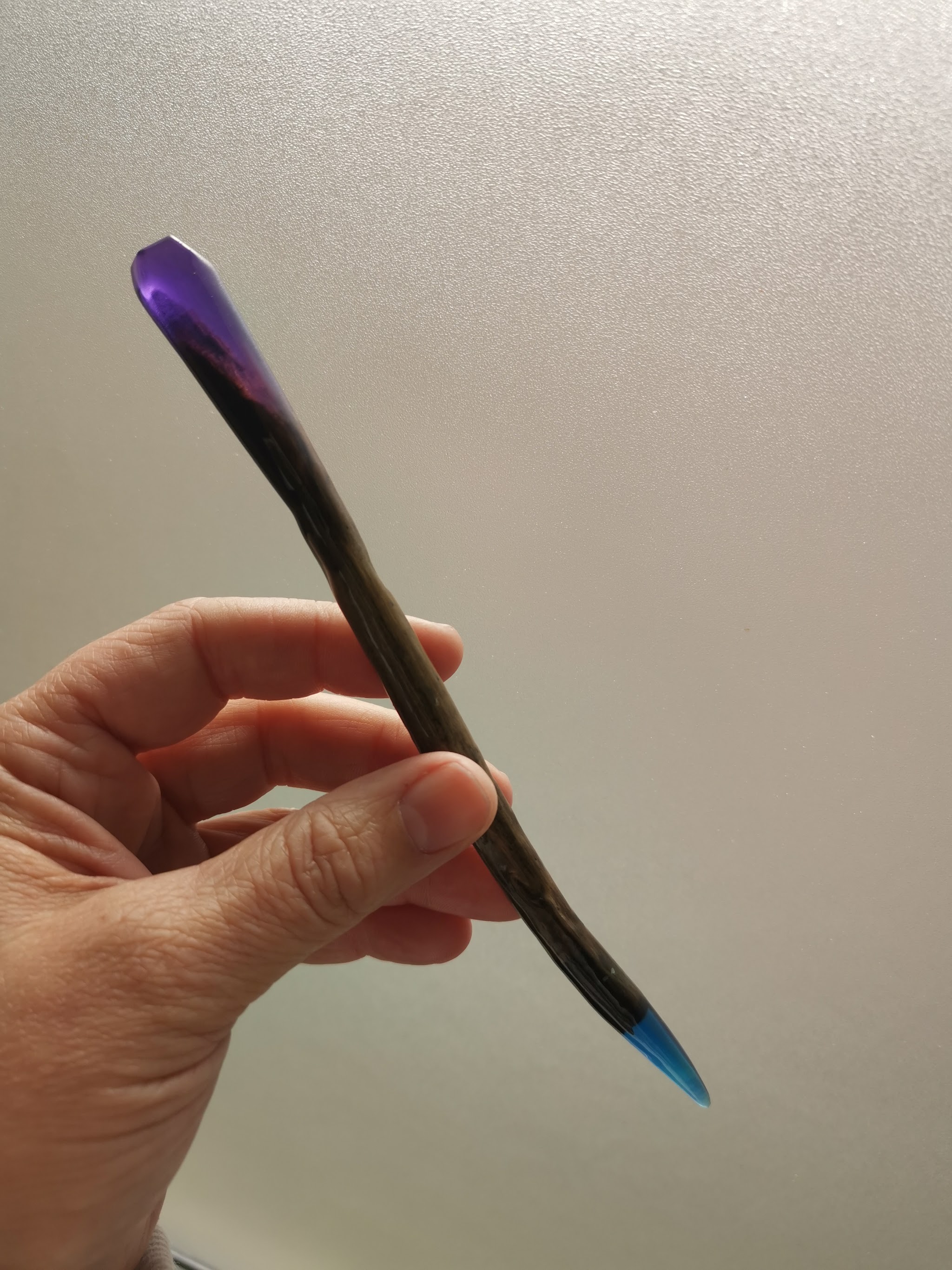 Drijfhout hairpin in de hand: paarse resin dop aan de bovenkant en blauwe resin punt aan de onderkant, verweerd hout daartussen