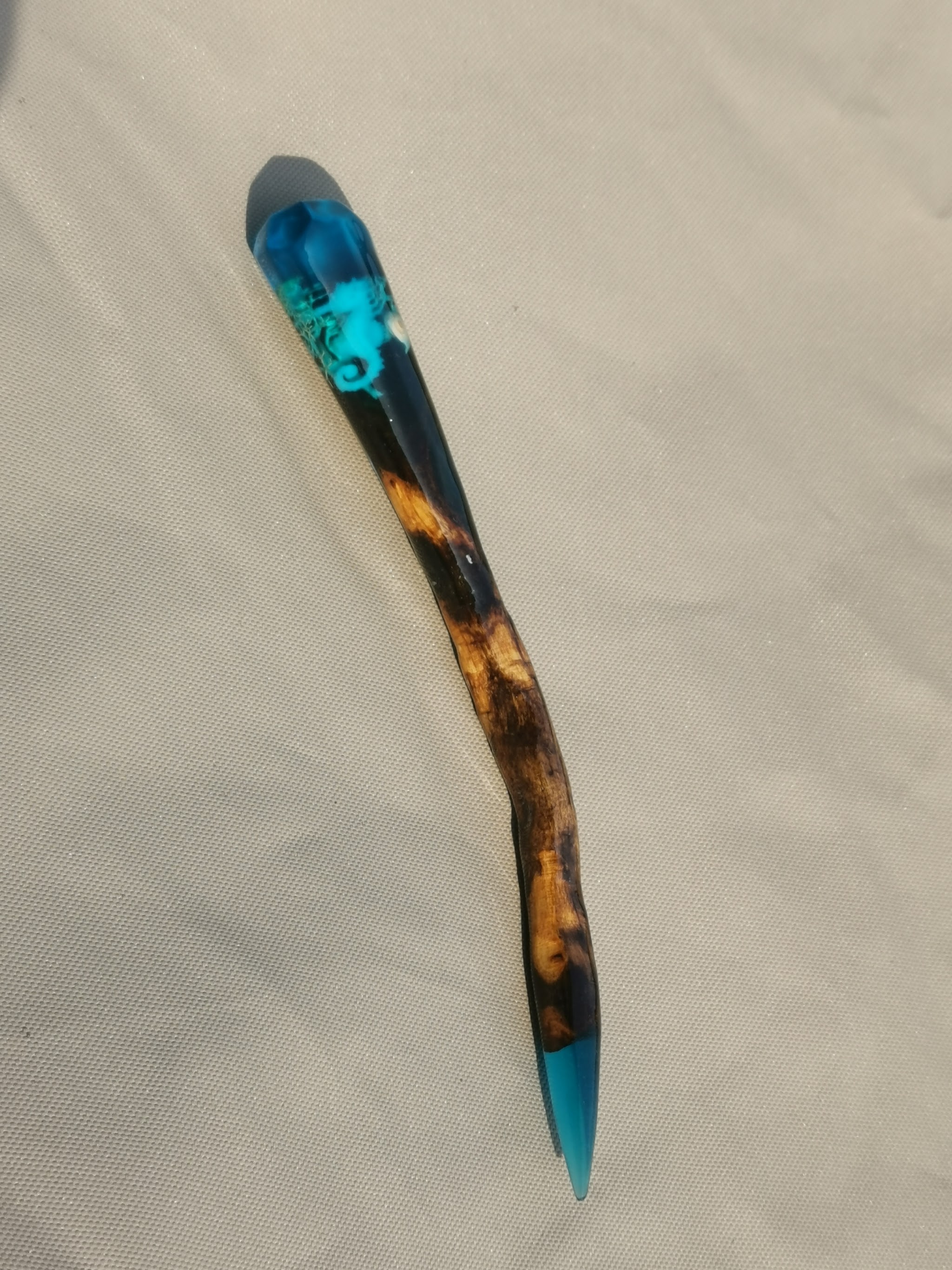 Afgewerkte drijfhout hairpin met turquoise resin dop met zeepaard als decoratie, liggend op een beige stof