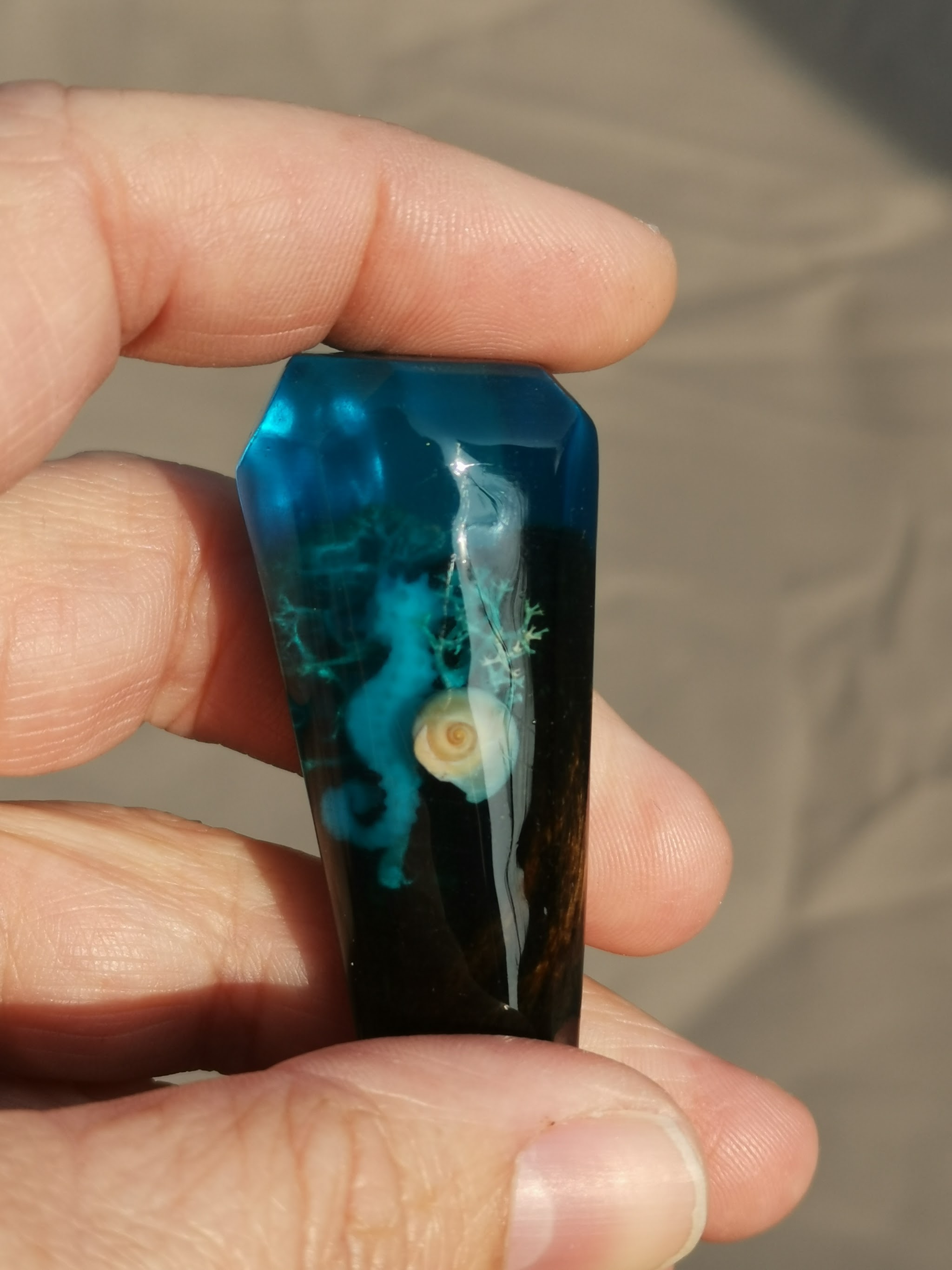 Close-up van de resin dop van een hairpin, vastgehouden tussen twee handen: zeepaard en schelp ingegoten als decoratie in turquoise en zwart hars