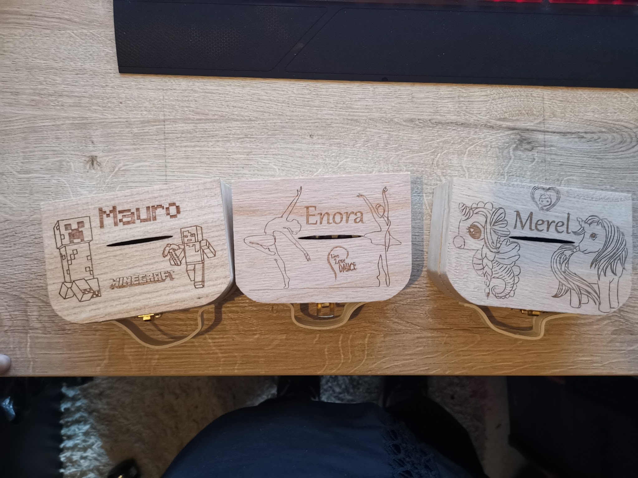 Overzicht van drie ronde houten doosjes op houten tafel, elk gegraveerd met een andere naam en persoonlijk motief